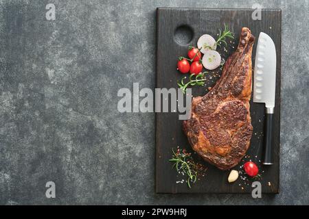 Tomahawk steak. Sliced grilled tomahawk beef steak with baked cherry tomatoes, herbs and salt on old wooden background. Preparing to grill.  Top view Stock Photo