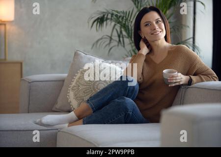 Happy dreamy middle aged woman sitting on comfortable sofa in living room with cup of black tea or coffee. Peaceful 30s lady enjoying no stress calm p Stock Photo