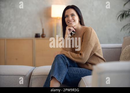 Happy dreamy middle aged woman sitting on sofa at home and looking at camera. Portrait of beautiful lady smiling and relaxing during autumn. Stock Photo