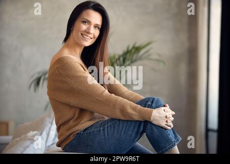 Happy dreamy middle aged woman sitting on sofa at home and looking at camera. Portrait of beautiful lady smiling and relaxing during autumn. Stock Photo