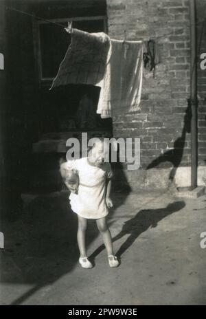 England.1930s. A charming photograph depicting an infant child. The young girl, who is wearing a white dress and sandals is standing in a backyard, clutching a soft toy under her arm and laughing. Towels and clothing are drying on a washing line above her head. Stock Photo