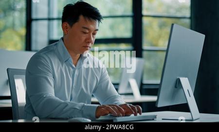 Portrait concentrated male office worker web-designer asian middle-aged man businessman sit at workplace use desktop computer developing tech program Stock Photo