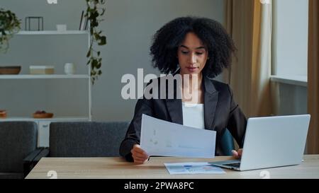 African teacher worker business woman work with graphics documents at home office check documentation enter financial data to laptop processing online Stock Photo