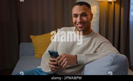 African american man sit on sofa in living room using online mobile application ethnic man playing game on phone internet communication with friends Stock Photo