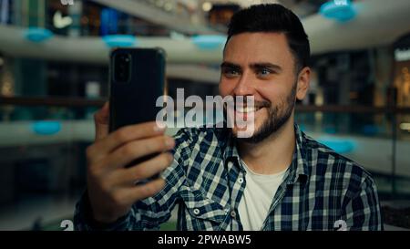 Smiling arabian caucasian male blogger businessman look at mobile phone camera talk video call in shopping mall happy young man chatting online Stock Photo