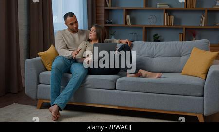 Diverse multiracial couple sitting on sofa surfing online website on laptop using computer at home relaxing woman and man talking buying retail goods Stock Photo