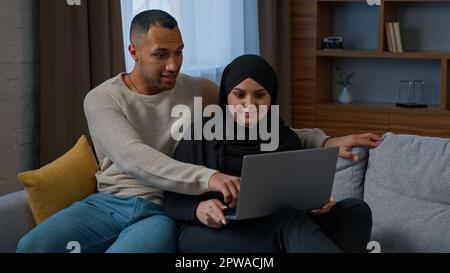 Married couple sit on sofa use laptop device choosing new apartment on internet website multiracial family shocked amazed diverse man woman search Stock Photo