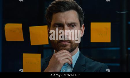 Thoughtful Caucasian businessman employee middle-aged man analyze information on stickers read sticky notes on glass wall standing in office planning Stock Photo