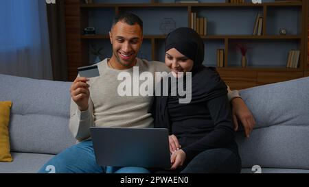 Multiracial couple sit on sofa use laptop purchasing on internet with credit card muslim wife woman in hijab and african american man husband shoppers Stock Photo