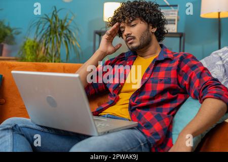Tired freelancer indian man use laptop, suffering from headache problem tension and migraine, stress at home room. Hindu guy works on notebook, sends messages, makes online purchases, watching movies Stock Photo
