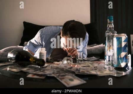 Young addicted junkie man sniffing snorting cocaine lines on mirror table with a lot of scattered money, alcohol, cards and pills through rolled bankn Stock Photo