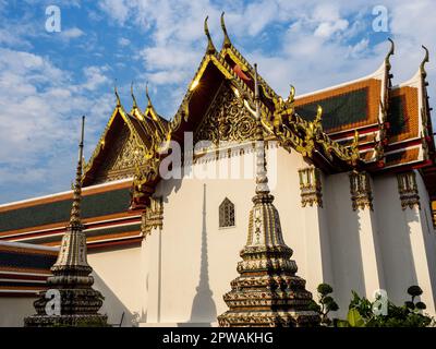 A section of the Wat Pho Temple, with intricate golden roof details reflecting the sun's rays. Two small spires with shadows are visible on the wall.
