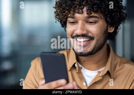 Young hispanic man close-up looking at phone screen, man at dinner reading online news from smartphone, businessman at workplace inside office browsing social networks happily. Stock Photo