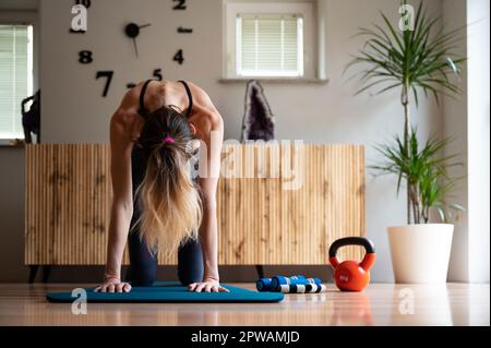 Front view of a slim fit young woman practicing yoga or pilates routine making a cat stretch exercise. On a workout mat in domestic living room. Stock Photo
