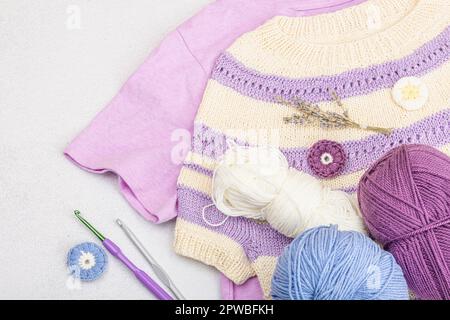 Handmade crocheted baby t-shirt in lilac tones. Stuff and props contains thread, hooks, knitting needles and craft decor. Light stone concrete backgro Stock Photo
