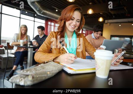Doing some studying on the go. A beautiful young university student using her smartphone in a coffeee shop while studying. Stock Photo