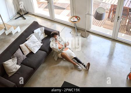 Happy young man with leg prosthesis sitting at home using phone, top view. Stock Photo