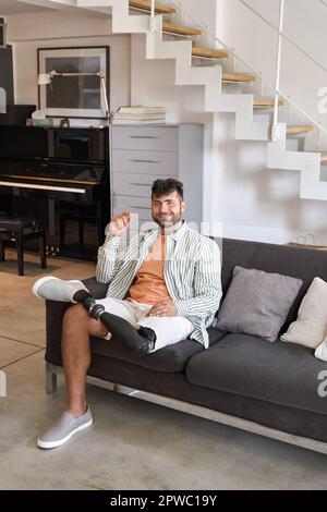 Happy young man with above knee leg prosthesis sitting on sofa at home. Stock Photo