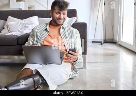 Man with disability leg prosthesis sitting home using phone and laptop. Stock Photo