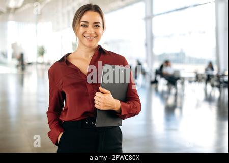 Portrait of a gorgeous successful confident positive blonde caucasian business lady dressed in an elegant wear, stands in a business center, holds documents, looks at camera, smiling friendly Stock Photo