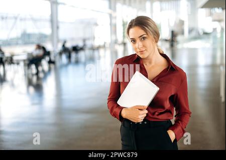 Portrait of a positive elegant beautiful confident successful blonde caucasian business lady dressed in a shirt, stands in a coworking center with a tablet in her hands, looks at the camera Stock Photo