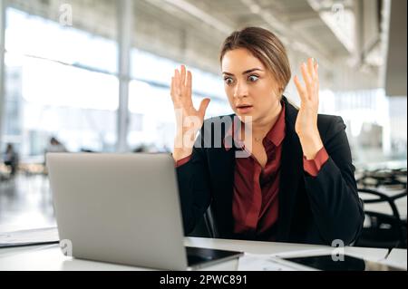 Shocked stunned caucasian woman, manager, office employee, sit in the office, looks in surprising at a laptop screen, reading unexpected news, gesticulates with hands, experiencing a misunderstanding Stock Photo