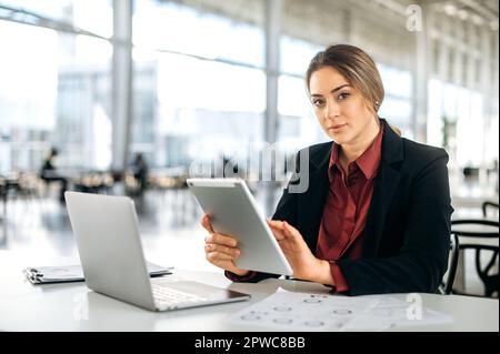 Serious beautiful elegant successful caucasian woman, entrepreneur, top manager, employee of marketing department, sits at a desk in the office, holds a tablet in her hands, looks at the camera Stock Photo