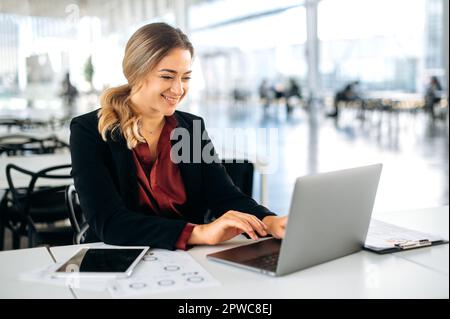 Confident successful positive caucasian business lady, executive, recruitment, product manager, sits in a business center, works in a laptop, looks at computer screen, smile, satisfied with the result Stock Photo