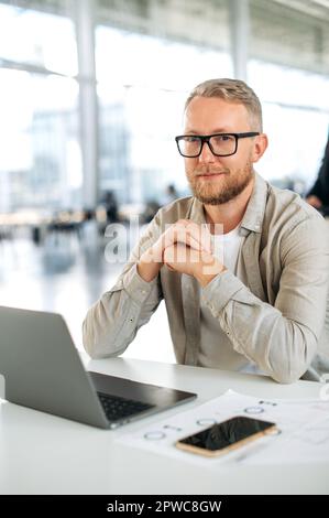 Vertical photo of a positive attractive clever caucasian business man, with glasses, programmer, company seo, software administrator, sits at a desk with a laptop in the office, looks at camera, smile Stock Photo