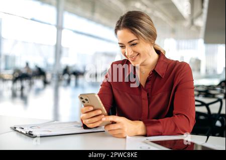 Positive attractive blonde caucasian business woman, office employee, manager, sit at her work desk in the office, using her mobile phone, messaging with colleague or client, reading news, smiling Stock Photo