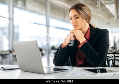 Serious focused elegant successful caucasian woman, entrepreneur, top manager, employee of marketing department, sits at a desk in the office, focused looks at laptop screen, working on a project Stock Photo