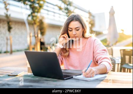 Concentrated lovely busy clever caucasian blonde woman, freelancer, student, marketing specialist, sits outdoors at a desk with a laptop, working on a project, takes notes, browsing information Stock Photo
