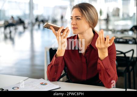 Focused busy successful confident caucasian business lady, sits at desk in office, using her smartphone, recording a voice message to a client, talking on speakerphone with a colleague, looking away Stock Photo