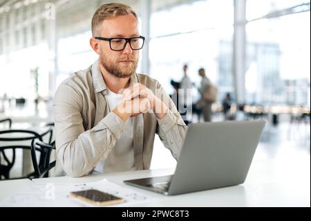 Thoughtful busy caucasian man with glasses, in casual wear, IT specialist, software tester, programmer, digital developer, works in a laptop in modern office, testing a new program, thinking Stock Photo