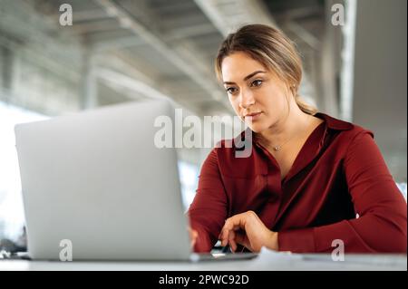 Focused busy confident caucasian woman, business lady, sits at workplace in the office, working in a laptop on a new important project, planning business plan, concentrated looks at laptop screen Stock Photo