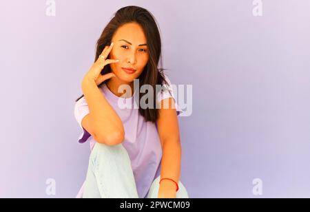 Full body of positive barefoot millennial Asian female in casual shirt and jeans sitting on floor in light violet studio Stock Photo
