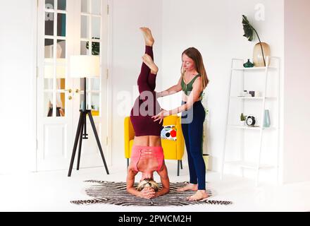 Full body adult woman supporting friend doing Headstand with Eagle Legs during yoga session in cozy room at home Stock Photo