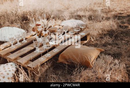 Set of elegant glassware placed on plank wooden table with floral arrangement on grassy meadow during festive autumn picnic Stock Photo