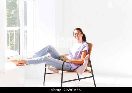 Full body of positive barefoot millennial Asian female in casual shirt and jeans sitting in light studio and drinking the coffee Stock Photo