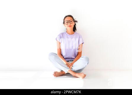 Full body of positive barefoot millennial Asian female in casual shirt and jeans sitting on floor in light studio Stock Photo