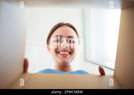 From below happy young female smiling and looking inside carton box while opening ordered goods Stock Photo