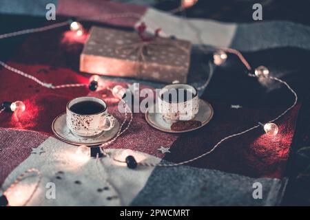 Cups of coffee and gift box placed on cozy bed with colorful checkered blanket and various pillows decorated with light garland and stars Stock Photo