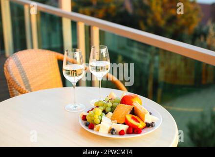 Assorted cheese cuts and fruit plate by two glasses of white wine on round table top standing on hotel balcony overlooking on summer forest at Stock Photo