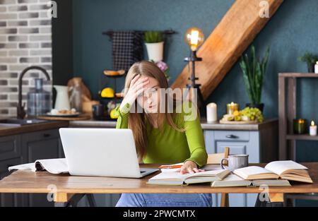 Young pensive woman freelancer sitting at table at home with laptop, making notes, staring thoughtfully into book, thinking about new ideas, female Stock Photo