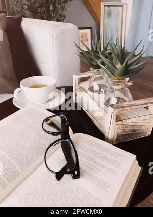 Eyeglasses on open book lying on round coffee table decorated with aloe plants on wooden stand next to cup of tea on saucer, living room interior Stock Photo
