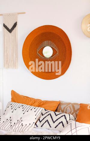 Patterned and colorful pillows on comfy bed in boho style bedroom with straw round carpet on white wall decorated with eye-shaped mirror next to Stock Photo