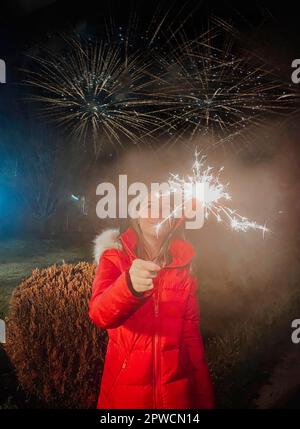 Happy teen girl holding sparkler while celebrating New Year outdoors in twilight during winter holidays, fireworks in sky on background Stock Photo