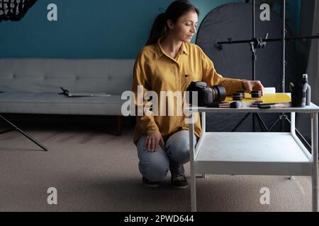 Joyful professional photographer lady preparing for shooting beauty product. Content creator using camera at home studio. Professional Photography Stock Photo