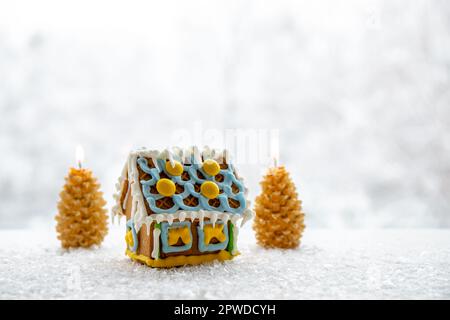 Cute little gingerbread house with, decorated with yellow and blue icing, spruce tree shape candles burning on background with winter landscape. Stock Photo