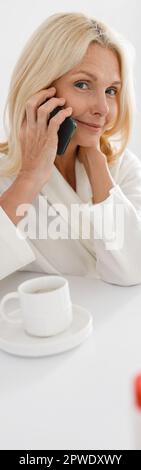 Vertical shot of senior lady speaking on smartphone sitting in modern cosmetology center Stock Photo